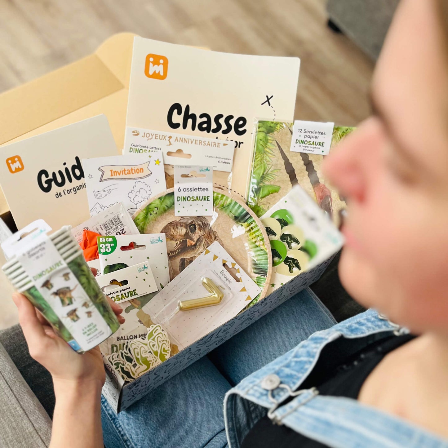 Personne ouvrant la box anniversaire Jimily sur le thème Dinosaure, découvrant le contenu : décorations, vaisselle jetable, bougie, chasse au trésor, guide pour les parents