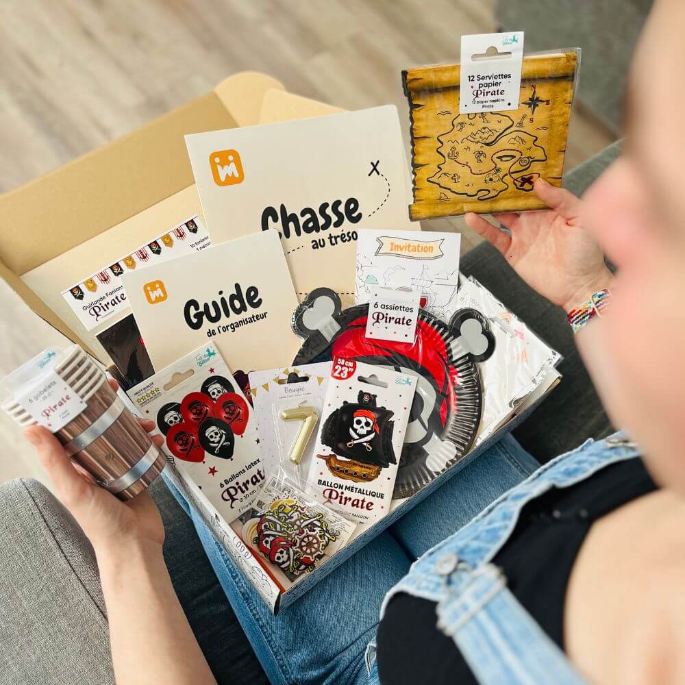 Personne ouvrant la box anniversaire Jimily sur le thème Pirate, découvrant le contenu : décorations, vaisselle jetable, bougie, chasse au trésor, guide pour les parents