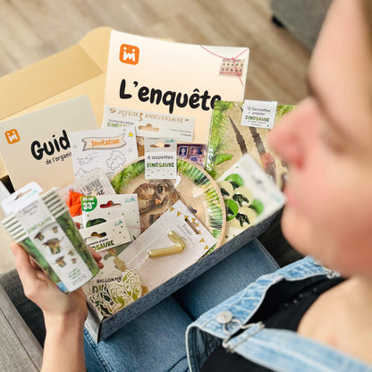 Personne ouvrant la box anniversaire Jimily sur le thème Dinosaure, découvrant le contenu : décorations, vaisselle jetable, bougie,  une enquête, guide pour les parents
